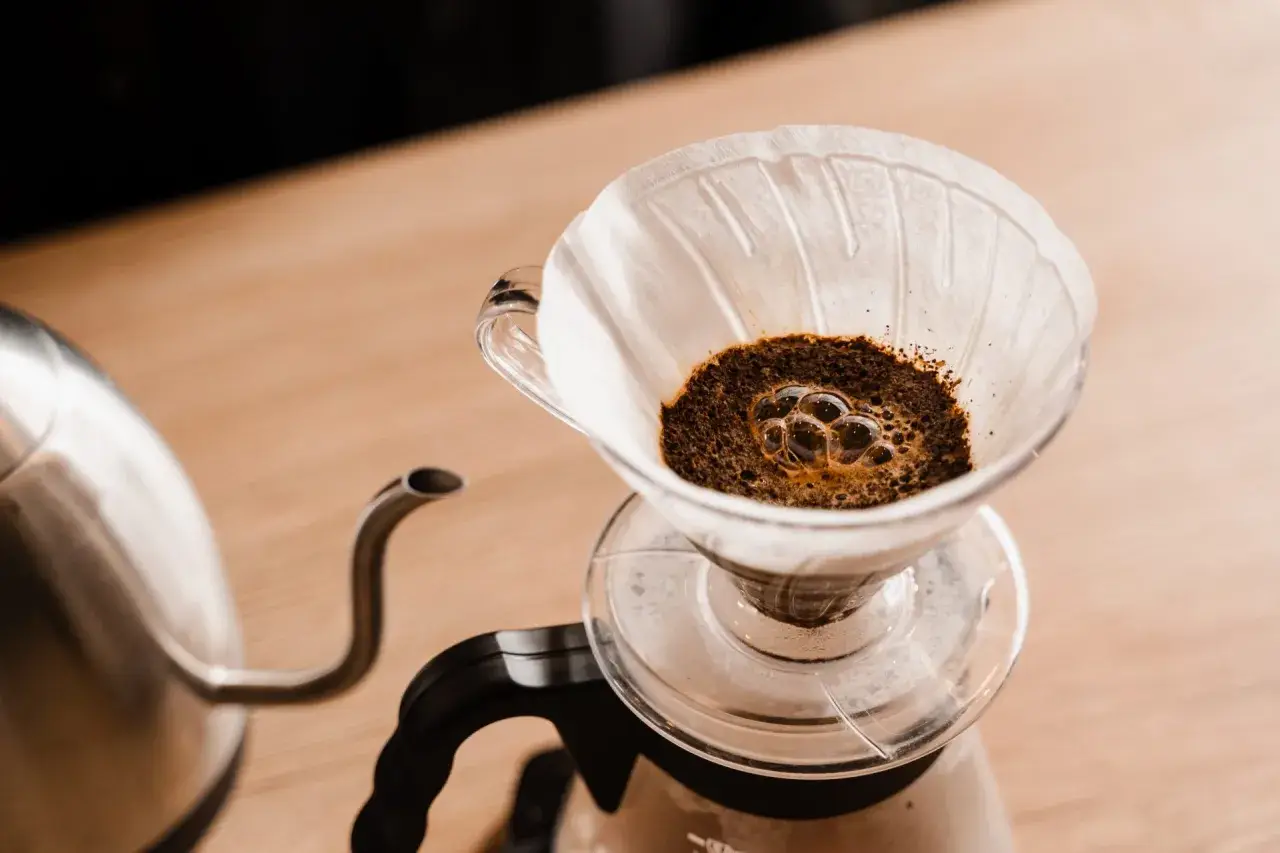 Frisch aufgebrühter Kaffee blubbert im Papierfilter. Ein Wasserkocher steht bereit, um das Ritual des Kaffeefilter selber machen zu beginnen.