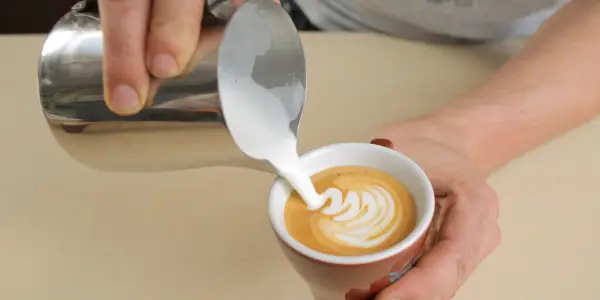 Barista gießt cremigen Milchschaum in eine Tasse Kaffee. Der Cappuccino Milchschaum bildet ein schönes Muster.