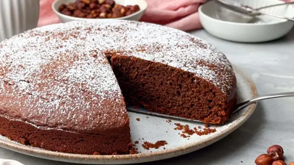 Ein saftiger Nutella Kuchen, bestäubt mit Puderzucker, wartet darauf, angeschnitten zu werden. Daneben eine Schüssel mit Haselnüssen.