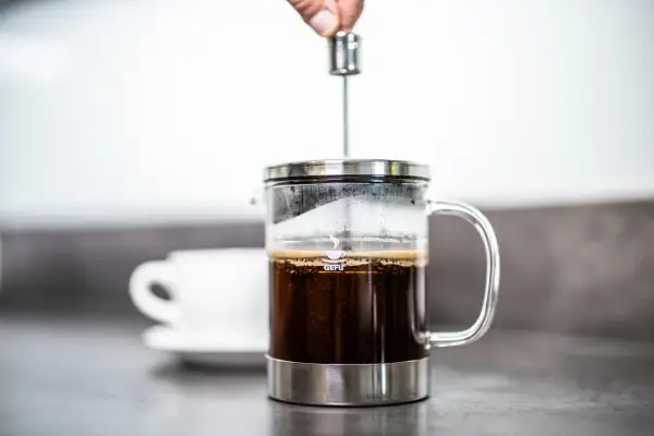 Eine Hand drückt den Stempel in eine French Press, um den perfekten Kaffee-Brew zuzubereiten. Eine Tasse wartet im Hintergrund.
